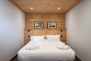 Una habitación de hotel con techo de madera, dos cuadros en la pared, una cama con ropa blanca y dos luces fijadas a la pared.