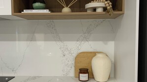 Een keuken met witte kastjes, een marmeren aanrechtblad, een houten plank met decoratieve objecten, en een potje met de tekst 'REFRESH'.