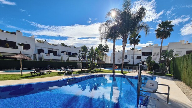 A swimming pool with lounge chairs and palm trees, surrounded by white buildings. - Cozy Beachside Haven (Pulpi)