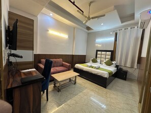 A modern hotel room with a bed, a desk, a chair, a ceiling fan, and a TV. - The Grand Palace  (Ghaziabad)