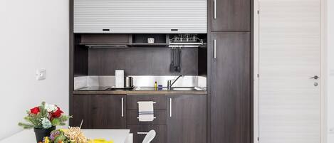 Una cocina compacta con armarios de madera oscura, encimera blanca y horno empotrado. Cuenta con un lavamanos pequeño y una ventana sobre la encimera.