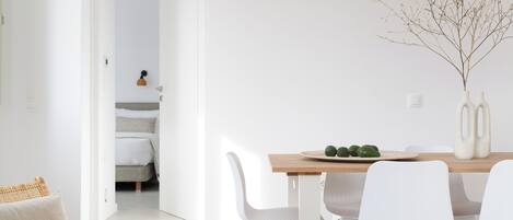 Un comedor moderno y minimalista con una mesa de madera y sillas blancas, un jarrón con ramas y vista a un dormitorio.