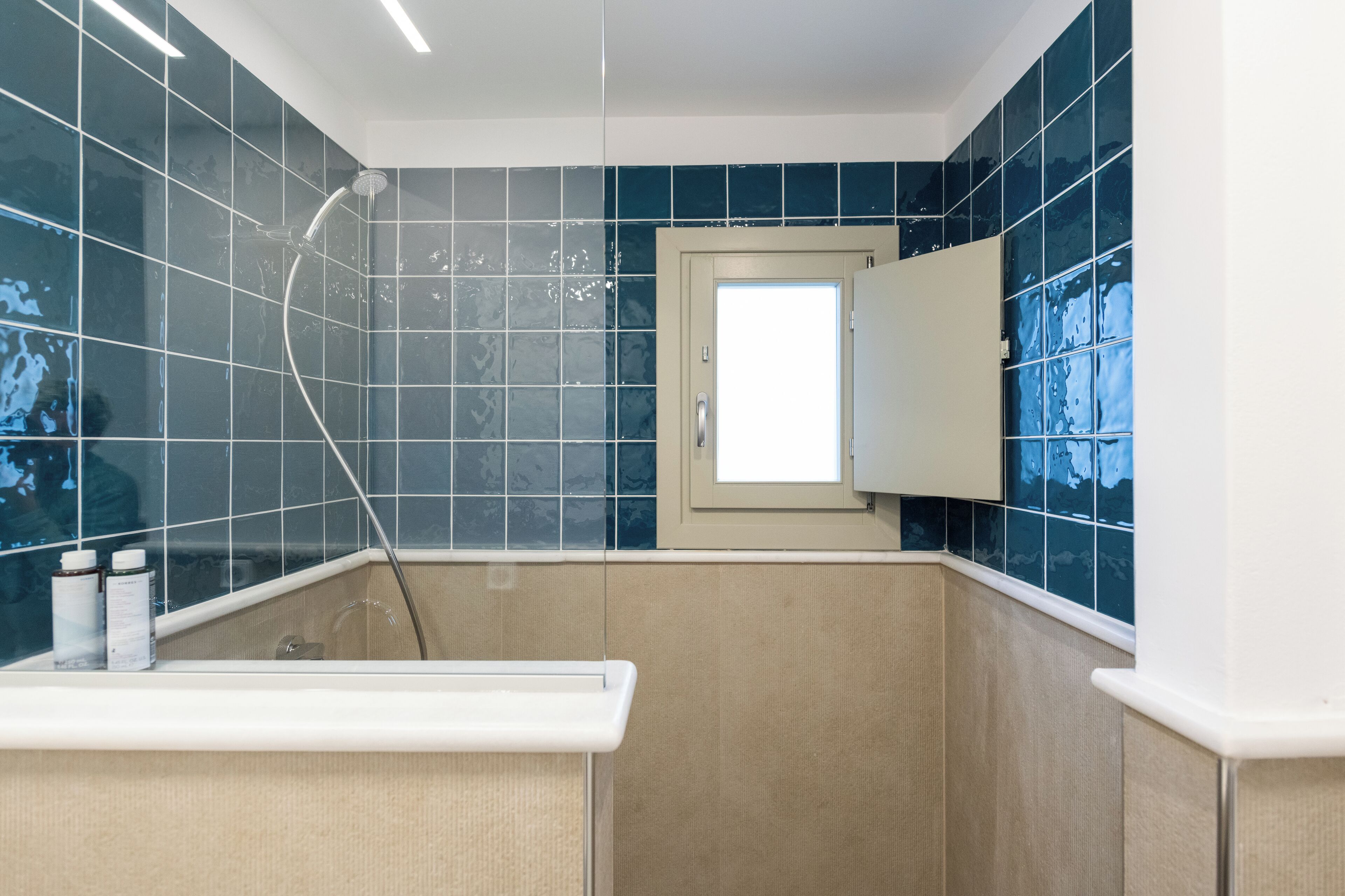 A bathroom with blue tiles, a shower, a sink with bottles, and a door with a window.