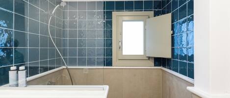 A bathroom with blue tiles, a shower, a sink with bottles, and a door with a window.