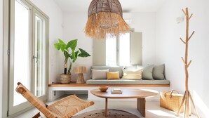 A modern living room with a sofa, a wooden coffee table, a wicker chair, and a large plant. - Triotissa Local Living Residences (Paros)