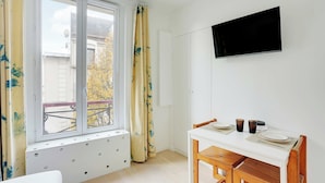 Una stanza con una finestra, una TV fissata al muro, un tavolo da pranzo con sedie e un piccolo armadietto bianco.