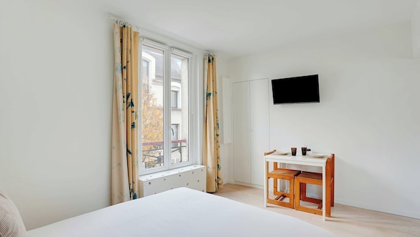 Una camera da letto con un letto, una scrivania con una sedia, una TV fissata al muro e una finestra con tende.