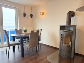 A dining area with a round table, four chairs, a view of the sea through a window, a wall-mounted light, and a wood-burning stove. - Apartment with a view of the ocean and a fireplace (Sassnitz)