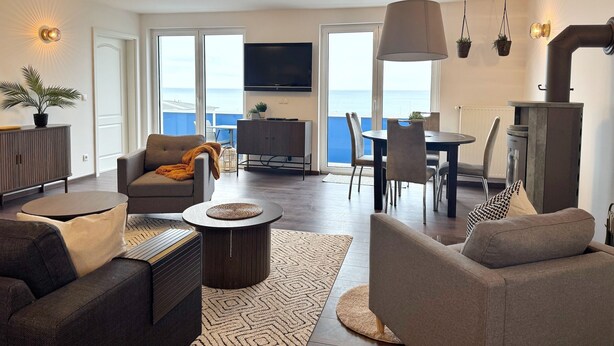 En moderne stue med sofa, lænestol og sofabord, et spiseområde med stole og bord, et tv ophængt på væggen og udsigt til havet gennem store vinduer.