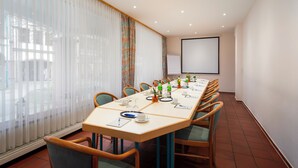 A conference room with a long table, chairs, a projector screen, and a large window with sheer curtains. - ARKADEN Hotel Kelkheim (Kelkheim)