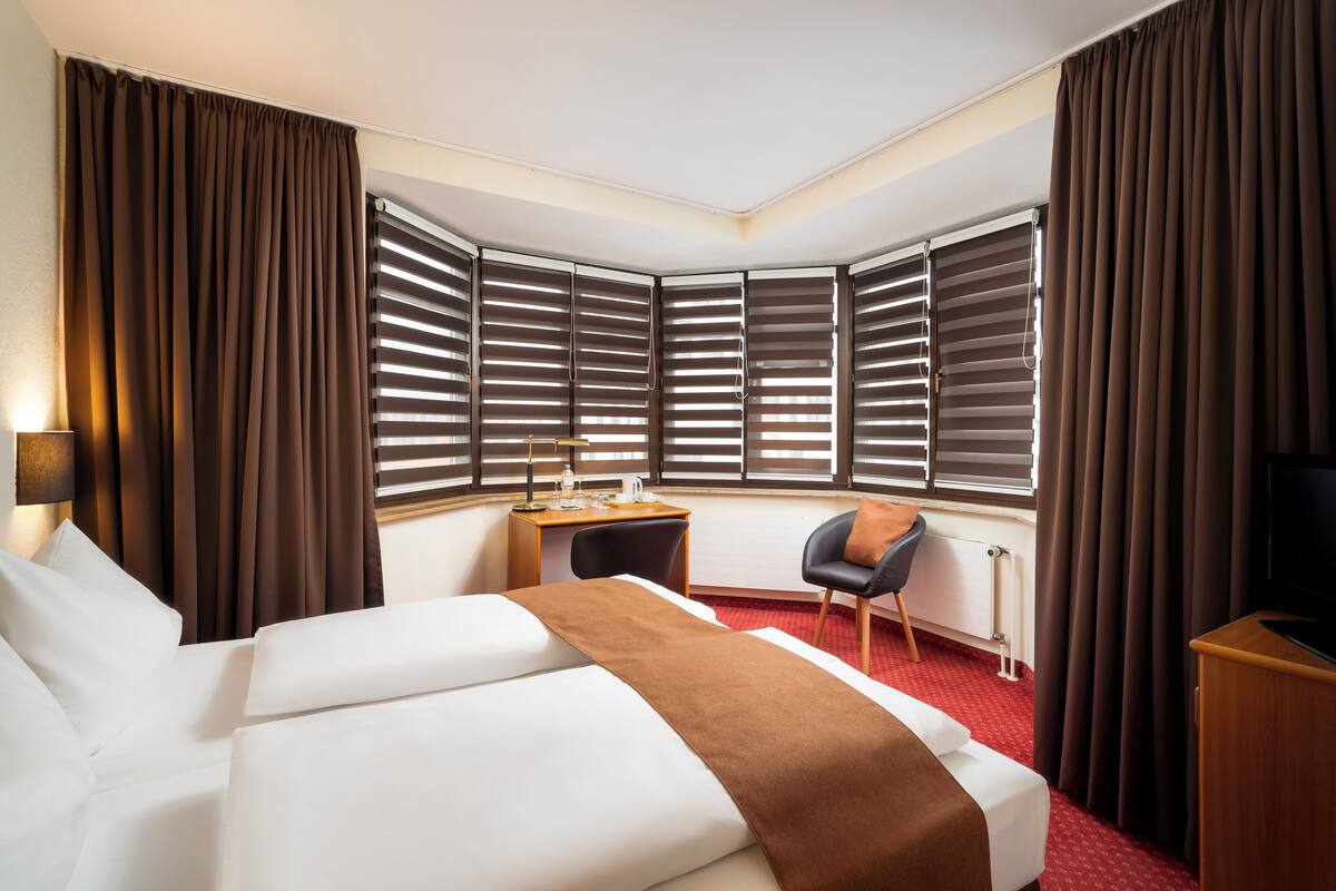 A hotel room with a bed, a desk, a chair, a TV, and large windows with blinds.