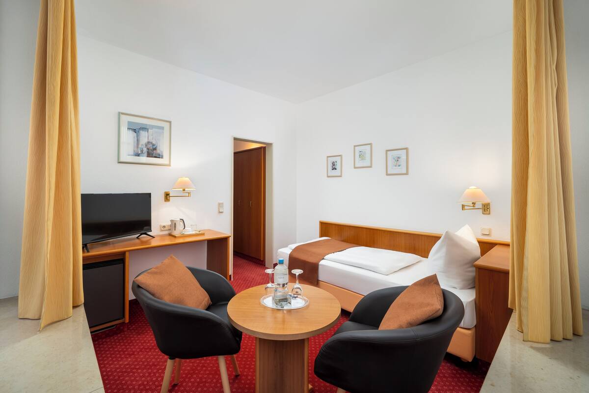 A hotel room with a bed, a desk with a computer, a small table with a bottle and glasses, two chairs, and a TV.