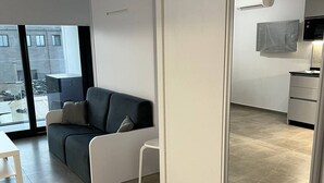 Een moderne woonkamer met een grijze bank, een klein wit tafeltje en een schuifdeur naar een keuken.