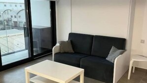 Een moderne woonkamer met een donkere bank, een witte salontafel en een uitzicht op een gebouw door grote ramen.