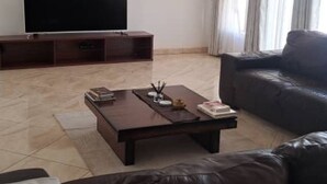 A living room with a flat-screen TV, a dark brown sofa, a coffee table, and a large window with curtains. - Gletwyn House Retreat in Harare (Harare)