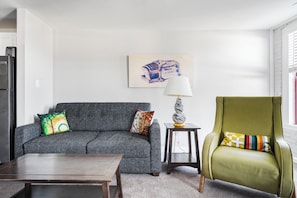 Un salon avec un canapé gris, un fauteuil vert, une table basse, une lampe et un tableau accroché au mur.