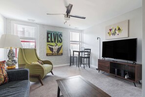 A living room with a green armchair, a sofa, a coffee table, a TV on a wooden stand, a ceiling fan, and a poster on the wall.