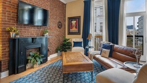 Una sala de estar con pared de ladrillo, un televisor de pantalla plana, chimenea, sofá, mesa de centro y vistas a edificios a través de amplios ventanales.