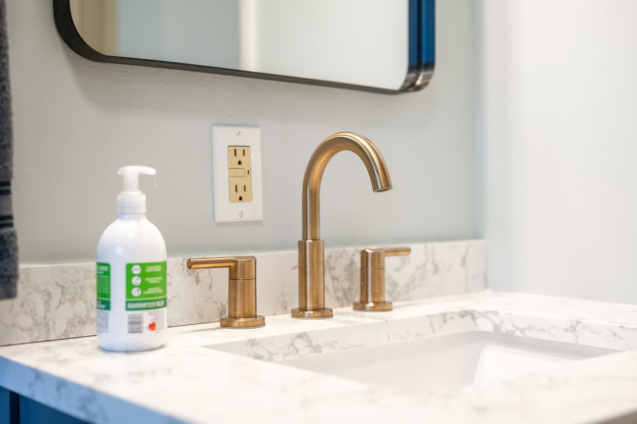 A bathroom with a marble sink, a gold faucet, and a bottle of hand sanitizer.