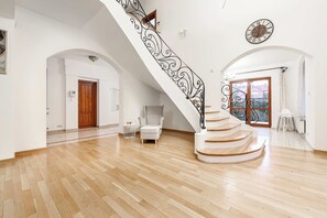 Una habitación amplia con piso de madera, una escalera blanca con barandal negro, una puerta de madera y un ventanal con vistas.