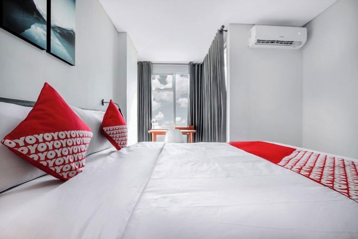 A hotel room with a large bed, red pillows, a desk, and a window with curtains.