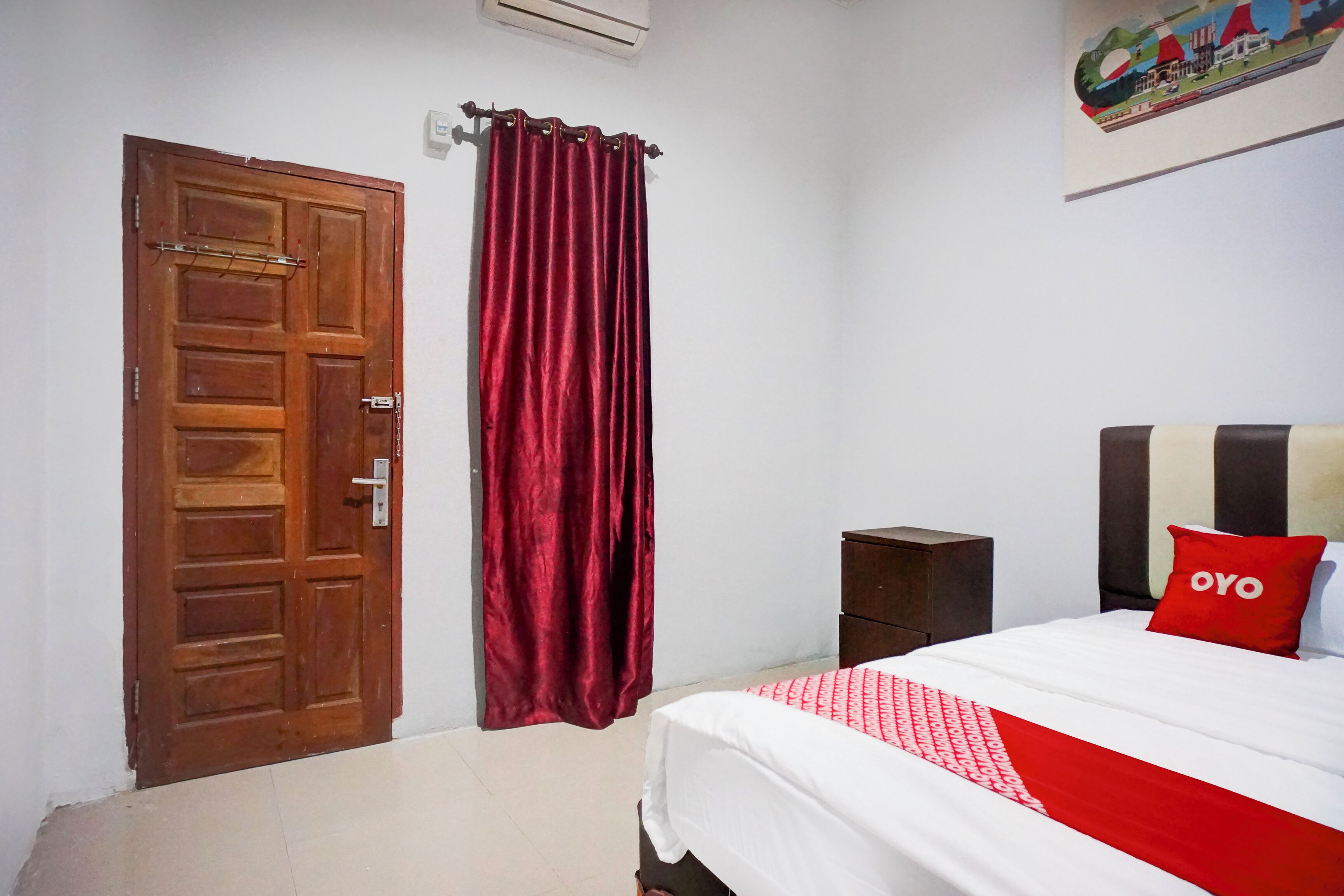 A bedroom with a wooden door, a red curtain, a bed with a white and red bedspread, and a small wooden nightstand.