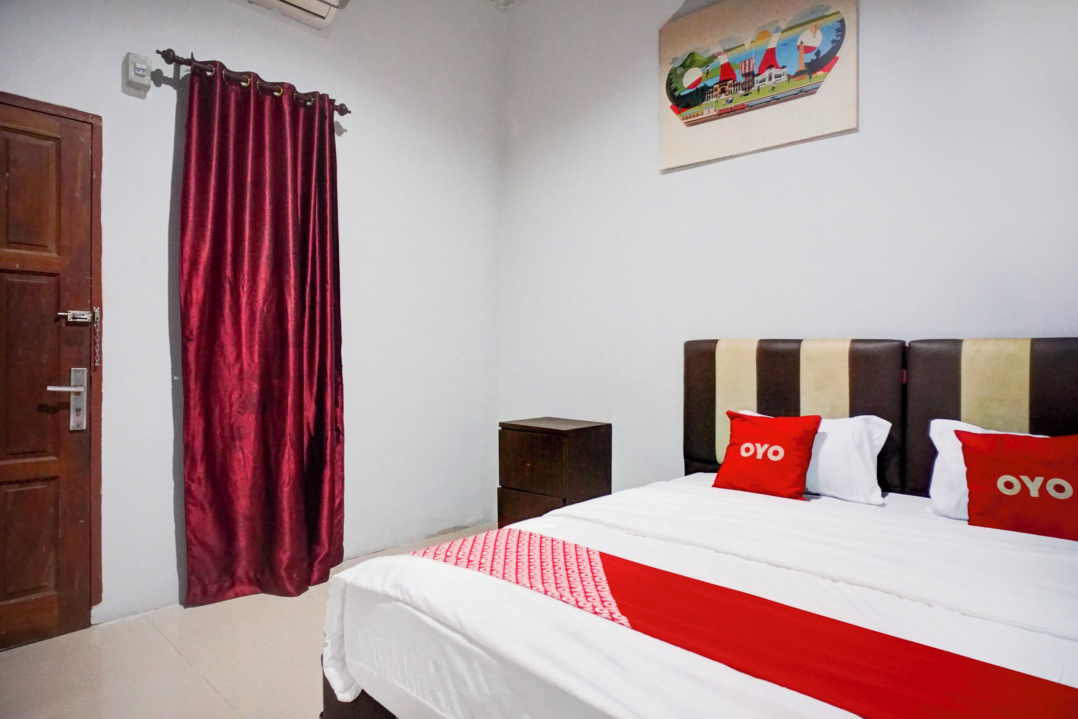 A bedroom with a bed, red and white bedding, a wooden door, a small wooden nightstand, and a colorful painting on the wall.