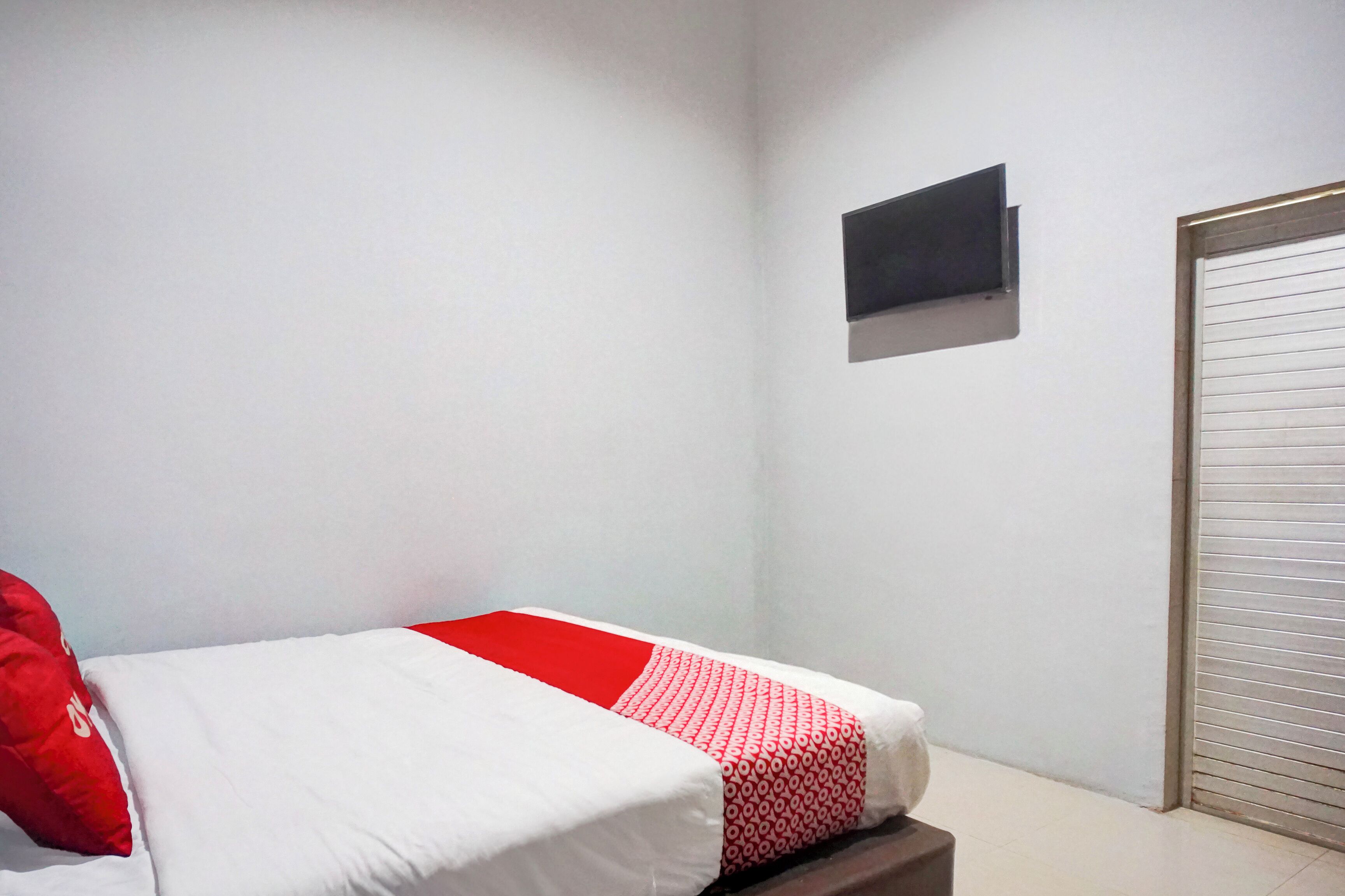 Une chambre à coucher avec un lit, une couvre-lit rouge et blanche, un oreiller rouge, un petit objet noir accroché au mur et une porte fermée.