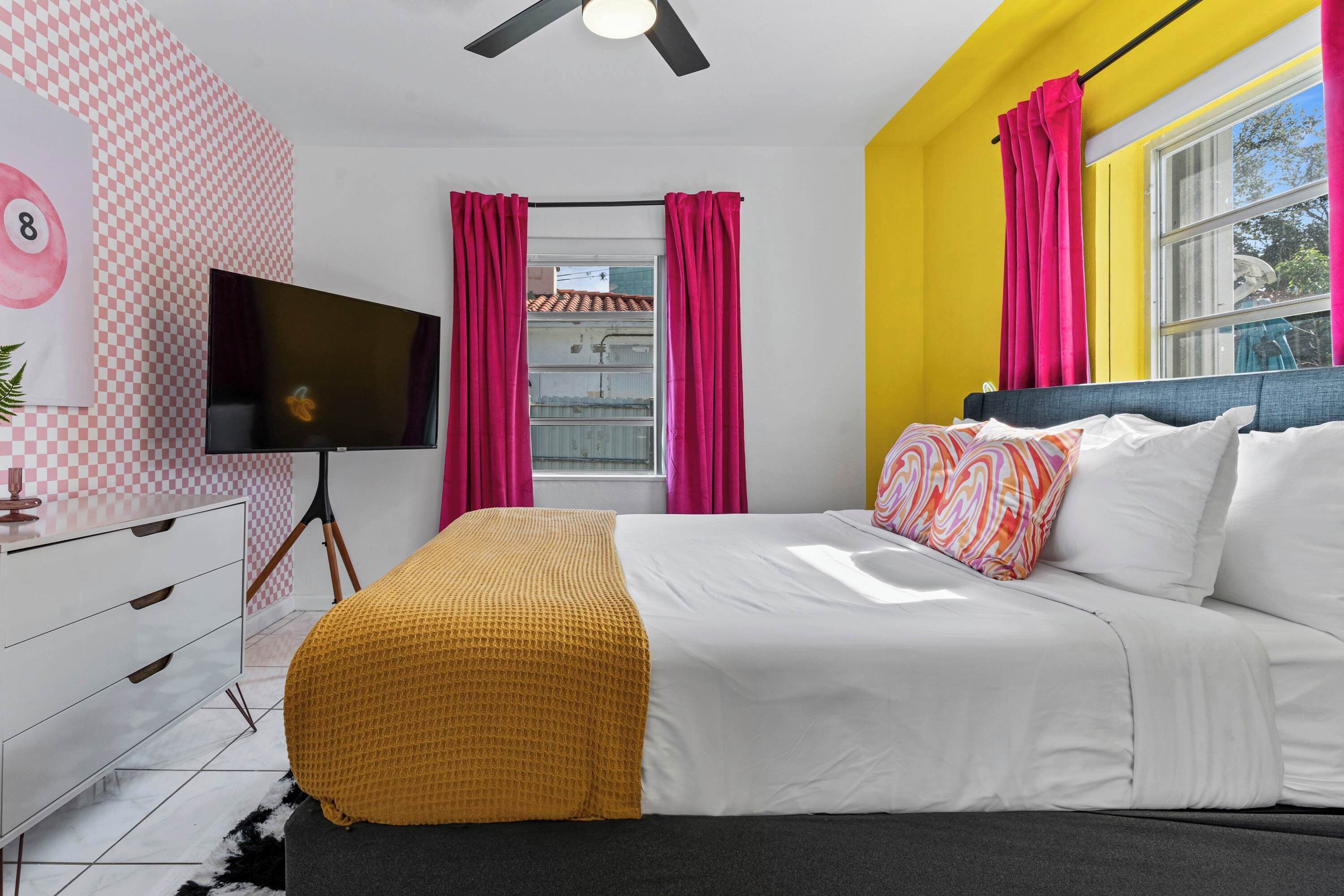 A bedroom with a bed, a TV, a window with curtains, and a wall with a checkered pattern.