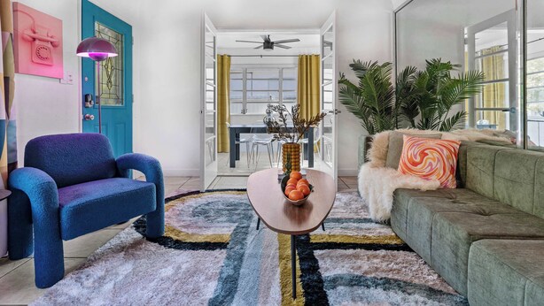 A living room with a blue armchair, a green sofa, a coffee table with a bowl of fruit, and a large window with curtains. - Retro Rewind, Stylish, Near Design District Miami (Miami)