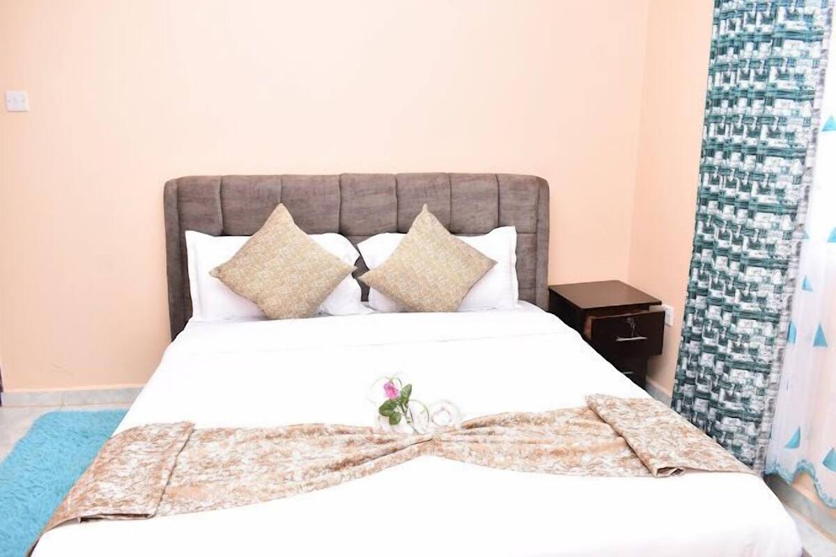 A neatly made bed with a floral bedspread and two decorative pillows, a nightstand with a drawer, and a patterned curtain.