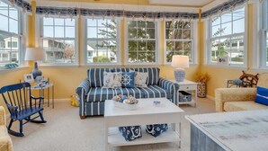 A cozy living room with a striped sofa, a coffee table, and a rocking chair. There are large windows with curtains, a ceiling fan, and a view of the outside. - Beachy Keen Steps to Sand, Miles from Stress (Avalon)