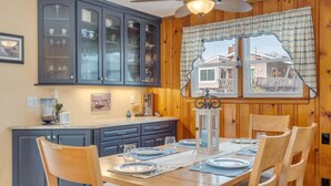 A dining area with a wooden table set for four, surrounded by dark blue cabinets with glass doors, a window with a view of a house, and a ceiling fan. - Beachy Keen Steps to Sand, Miles from Stress (Avalon)