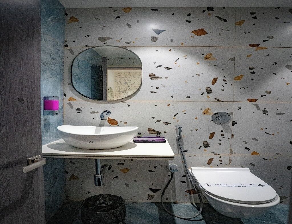 A modern bathroom with a white sink, a round mirror, and a toilet. The walls have a terrazzo finish with multicolored stone pieces.