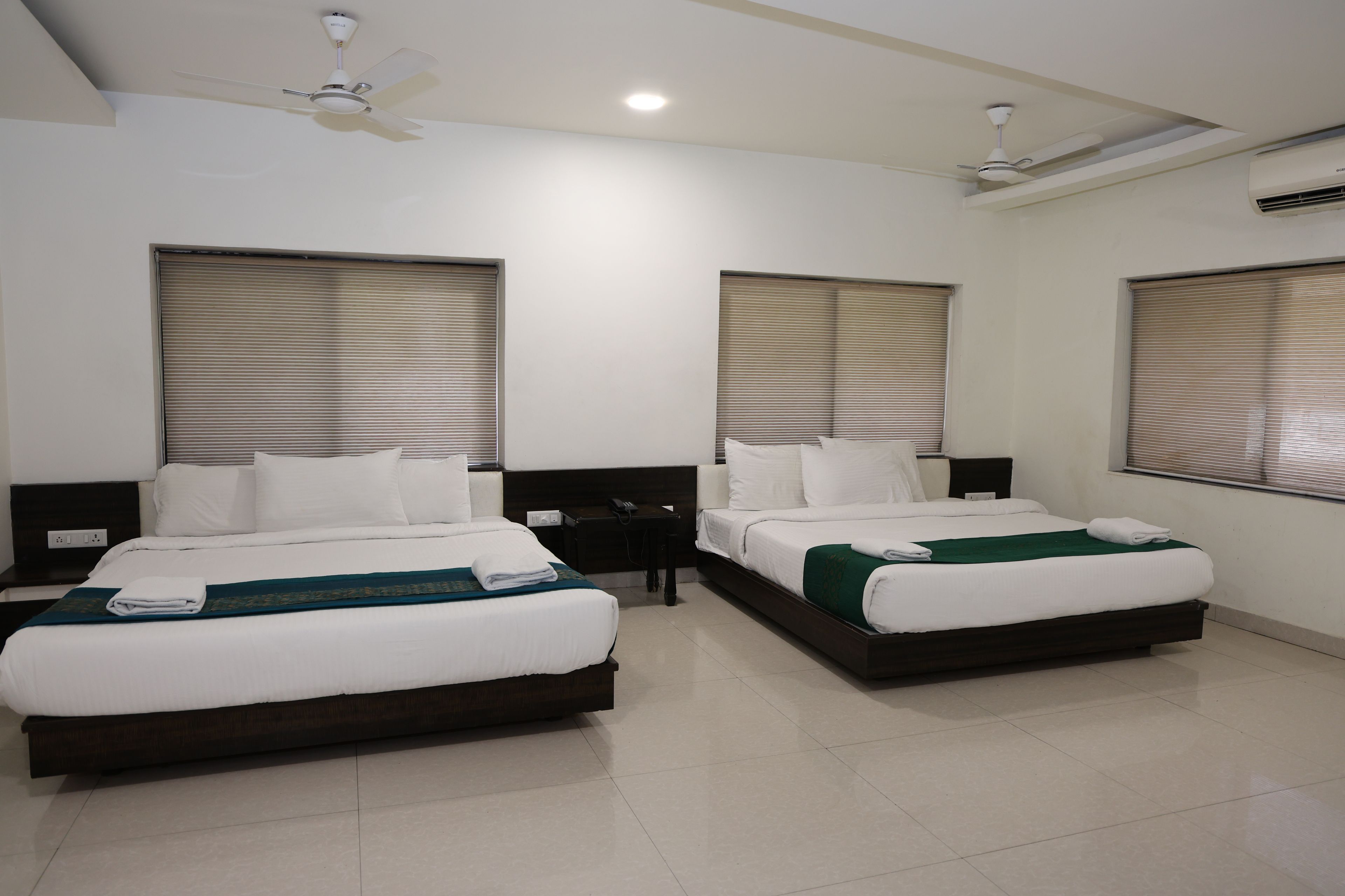 A hotel room with two single beds, a ceiling fan, an air conditioning unit, and a window with blinds.