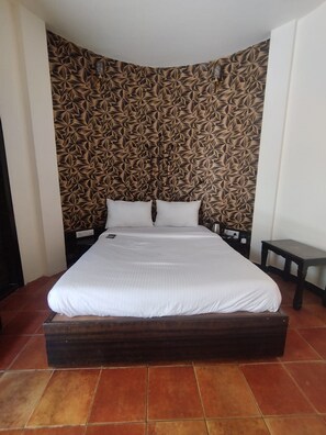 Una cama con colchón y almohadas blancas, cabecera de madera oscura y pared con un diseño detrás.