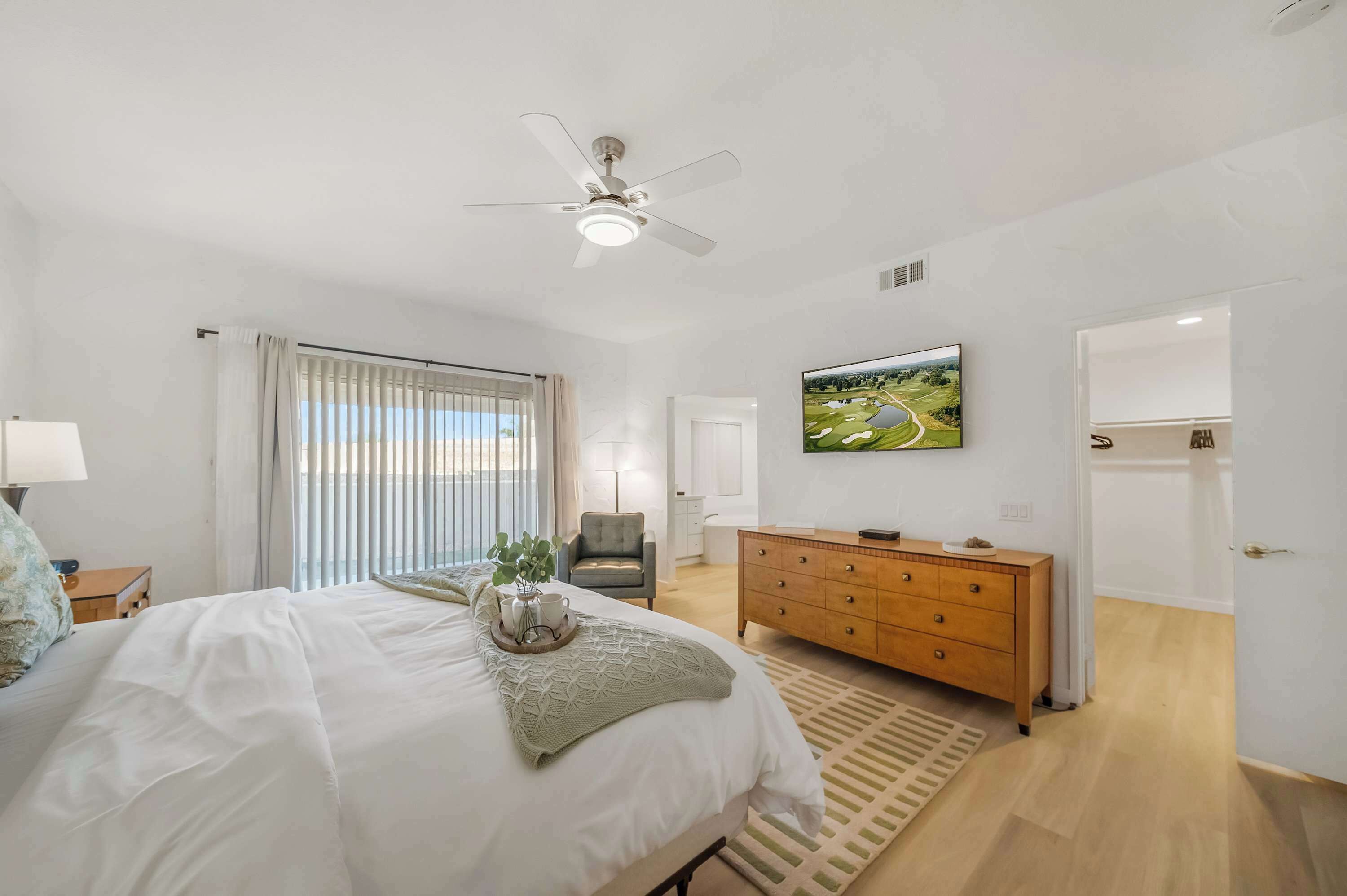 Une chambre à coucher comprenant un lit, un ventilateur de plafond, une télévision, une commode et une fenêtre avec des rideaux.