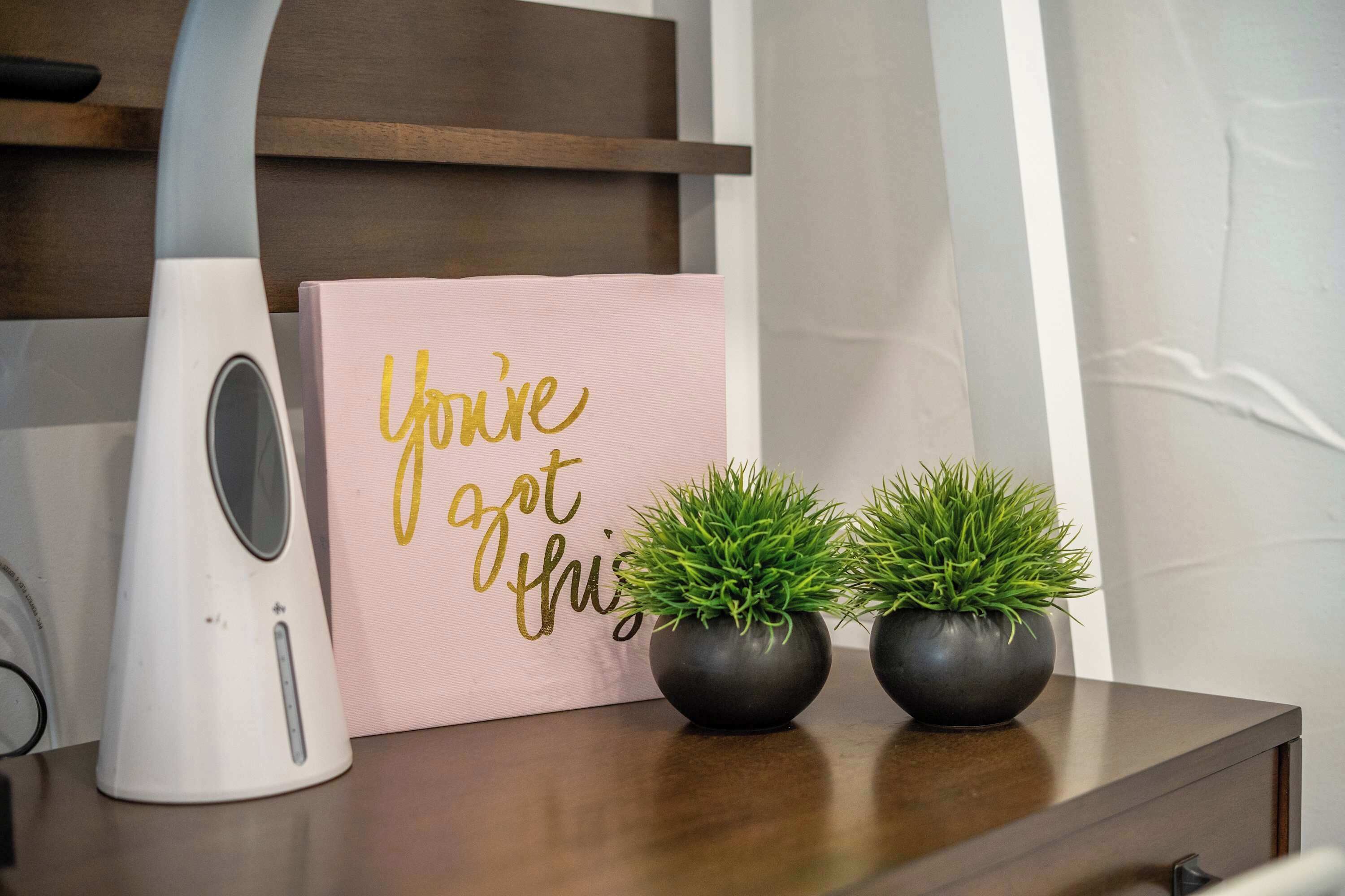 Une table de chevet avec une lampe blanche, un panneau rose avec des lettres dorées et deux plantes en pot.
