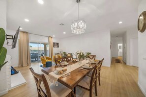 A dining area with a wooden table set for six, surrounded by wooden chairs. A chandelier hangs from the ceiling, and a large window with curtains is visible in the background. - Golf Resort Escape | Fairway Views + Pools & Pickleball (Palm Desert)