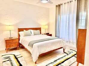 A bedroom with a wooden bed, two bedside tables with lamps, a patterned rug, and a window with curtains. - Golf Resort Escape | Fairway Views + Pools & Pickleball (Palm Desert)