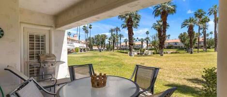 An outdoor patio with a round table and chairs, a grill, and a view of a well-maintained lawn with palm trees and buildings in the background.