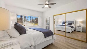 Un dormitorio con una cama grande, un ventilador de techo, un espejo amplio y una ventana con vistas a árboles.