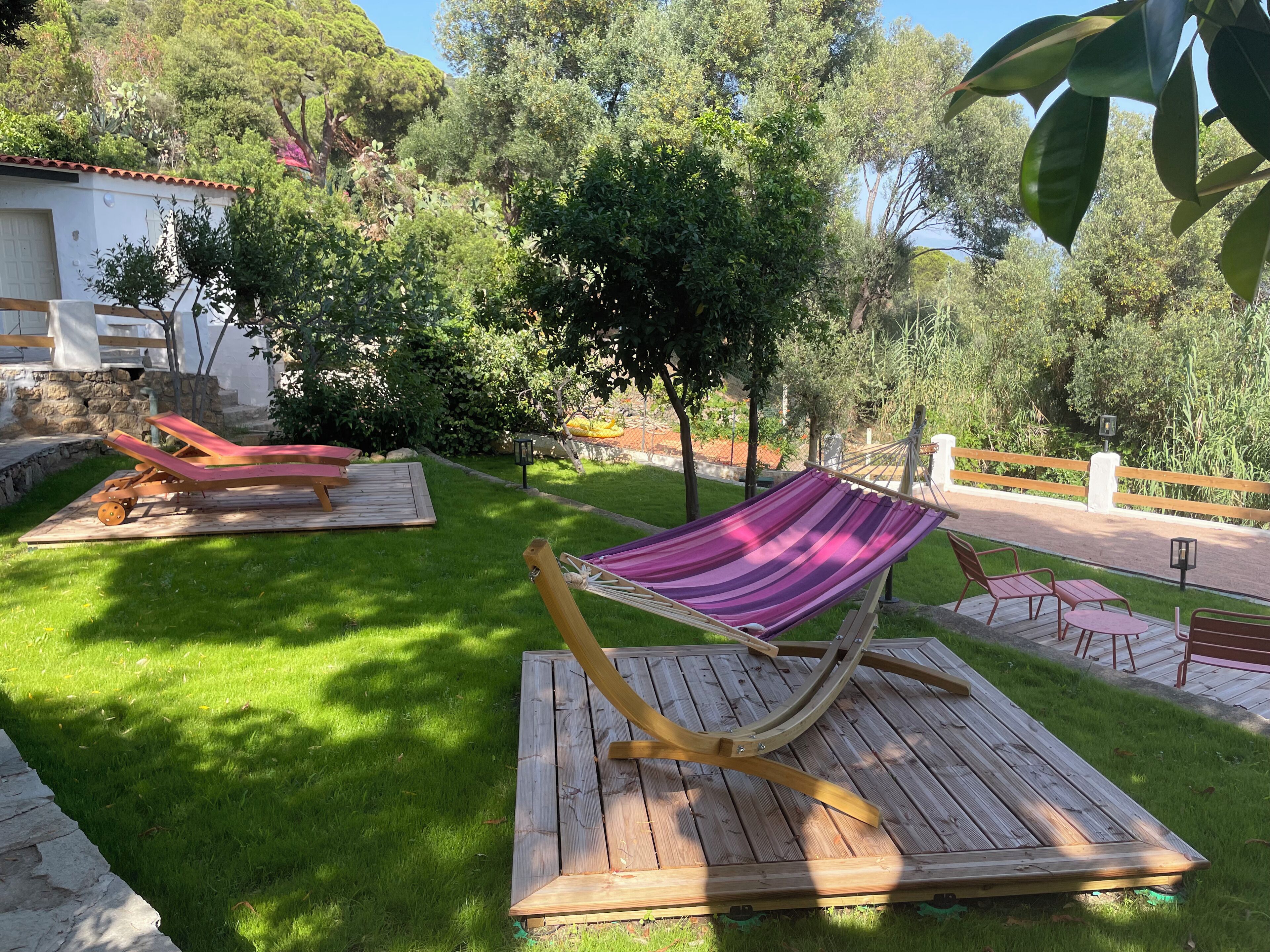 A hammock on a wooden platform in a garden with a stone wall and trees in the background.