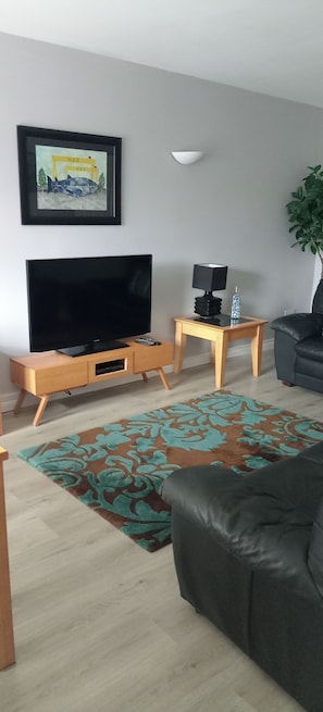 A living room with a flat-screen TV, wooden side table, lamp, and a patterned rug. - Bangor - 2 Bed Apartment- Sleeps 4 -privateparking (Bangor)