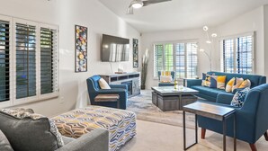Un vaste salon comprenant un canapé bleu, un fauteuil gris et un pouf à motifs. Un téléviseur à écran plat est fixé au mur, ainsi qu’un ventilateur de plafond et une grande fenêtre équipée de stores.