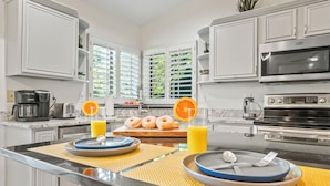 Une cuisine moderne avec des meubles blancs, un îlot central où sont disposés des aliments pour le petit-déjeuner, et une grande fenêtre équipée de stores.