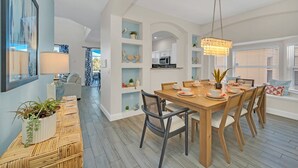 A dining area with a wooden table, chairs, and a chandelier. There are shelves with decorative items and a view of the kitchen in the background.