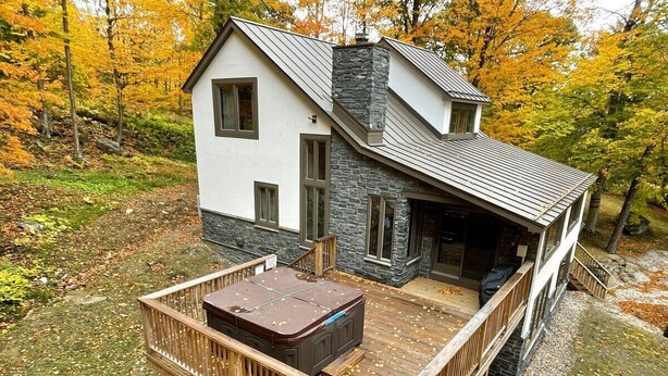 A cabin with a deck and a stone chimney. - The Navillus House: Hot Tub, Ski On/Ski Off, A/C. Discounted Ski Rentals. (Killington)