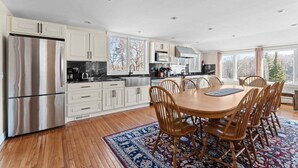 A modern kitchen with white cabinets, stainless steel appliances, and a dining area with wooden chairs and a patterned rug. - The Navillus House: Hot Tub, Ski On/Ski Off, A/C. Discounted Ski Rentals. (Killington)