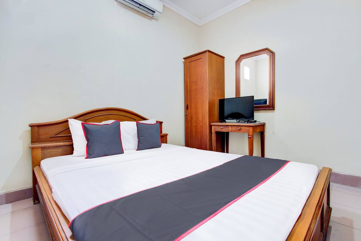 A neatly made bed with a white and black bedspread, a wooden headboard, and two pillows. There is a wooden wardrobe and a small desk with a computer monitor.